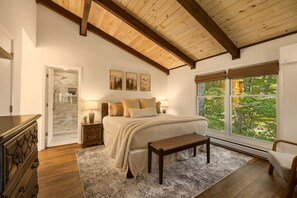 Ein Schlafzimmer mit einem großen Bett, einer Bank, einem Nachttisch, einer Kommode, einem Fenster mit Blick auf Grünflächen und einer Holzzieldecke.