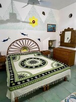 Un dormitorio con cama, cómoda, espejo y decoraciones en la pared, incluyendo un sol, una flor y peces.