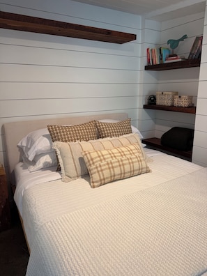 A bedroom with a bed, pillows, a wooden headboard, a shelf with books and decorative items, and a wall with horizontal lines.