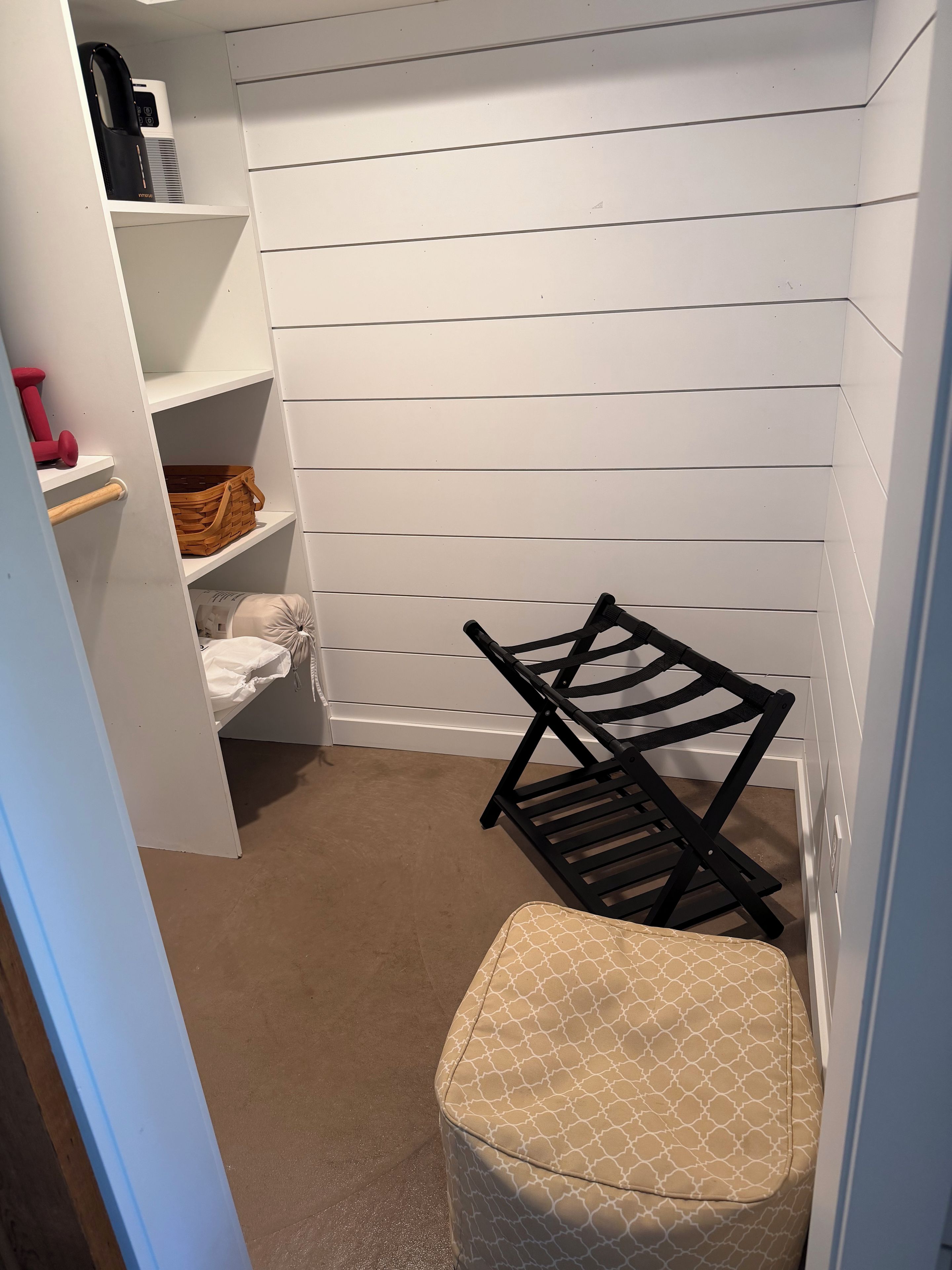 Un cuarto de lavandería con una silla negra, un banco blanco y estantes que contienen una cafetera, mancuernas y una cesta.