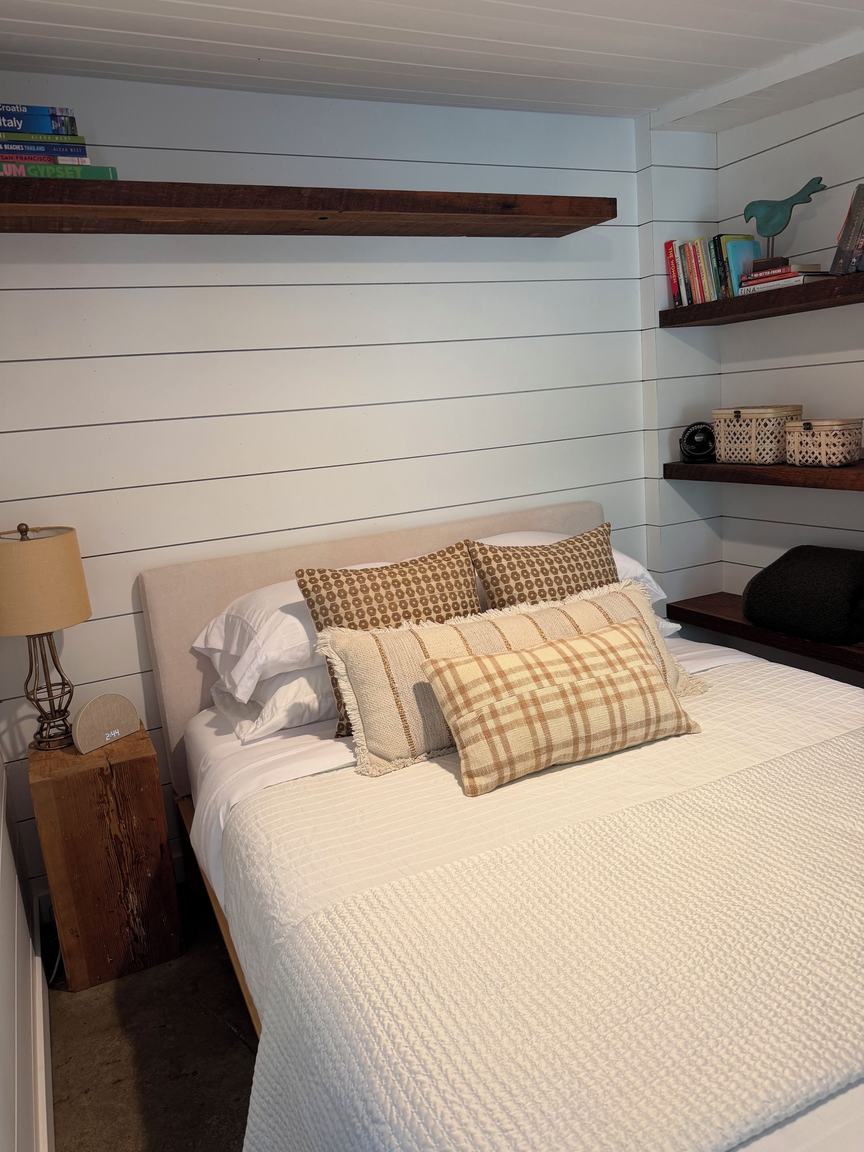 A bedroom with a bed, pillows, a nightstand, a lamp, a shelf with books, and a wall-mounted shelf with decorative items.