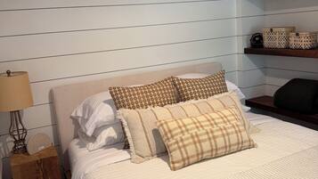 A bedroom with a bed, pillows, a nightstand, a lamp, a shelf with books, and a wall-mounted shelf with decorative items.