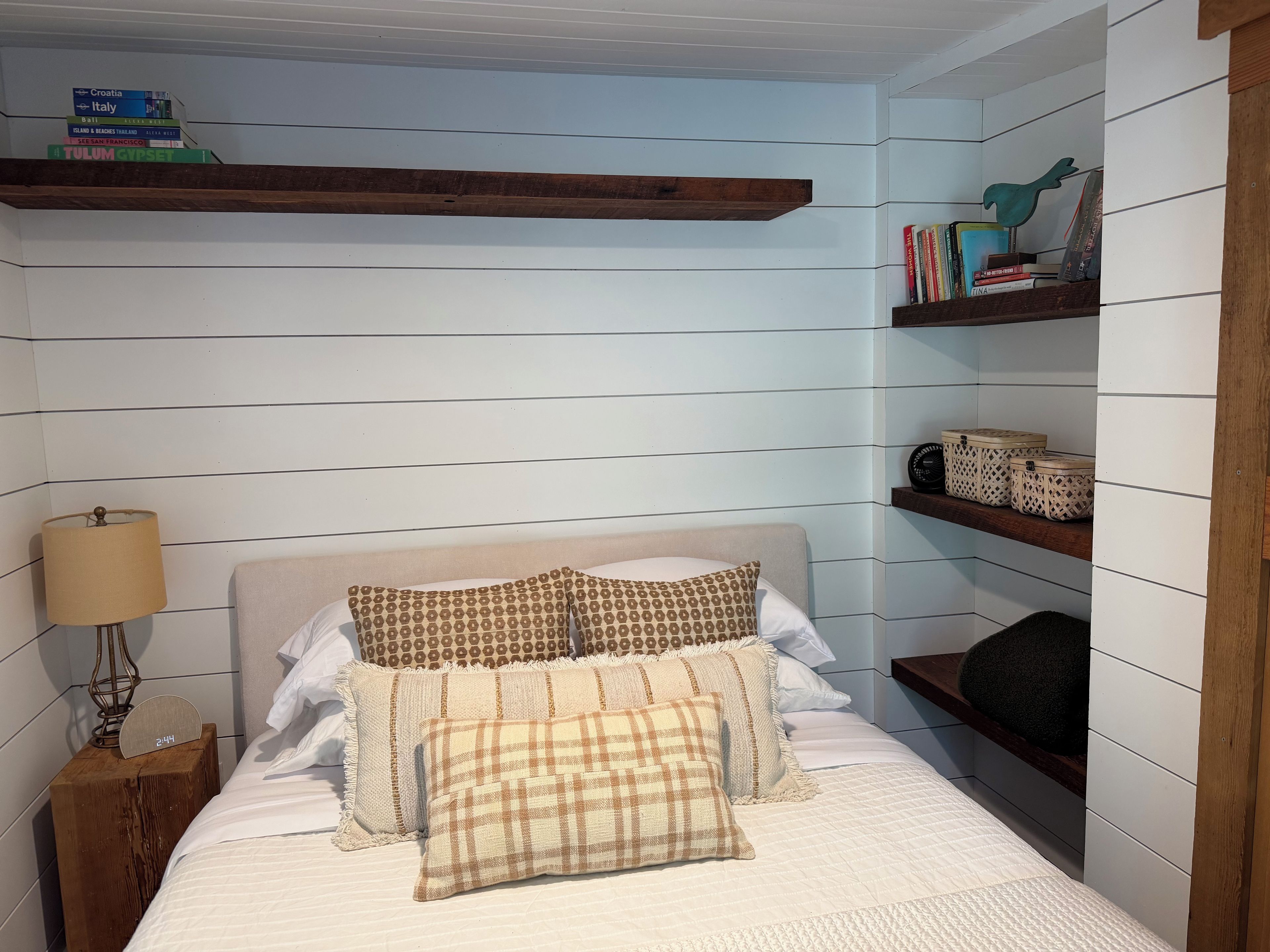 A bedroom with a bed, a nightstand, a lamp, a clock, and shelves with books and decorative items.