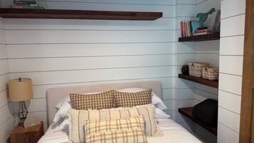 A bedroom with a bed, a nightstand, a lamp, a clock, and shelves with books and decorative items.