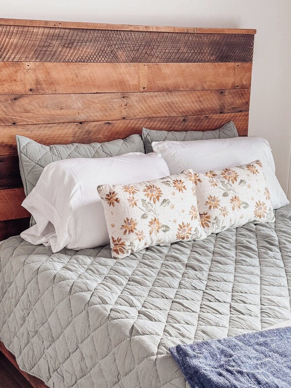 Una cama con una colcha gris acolchada, cabecera de madera y dos almohadas, una blanca lisa y otra con estampado floral.
