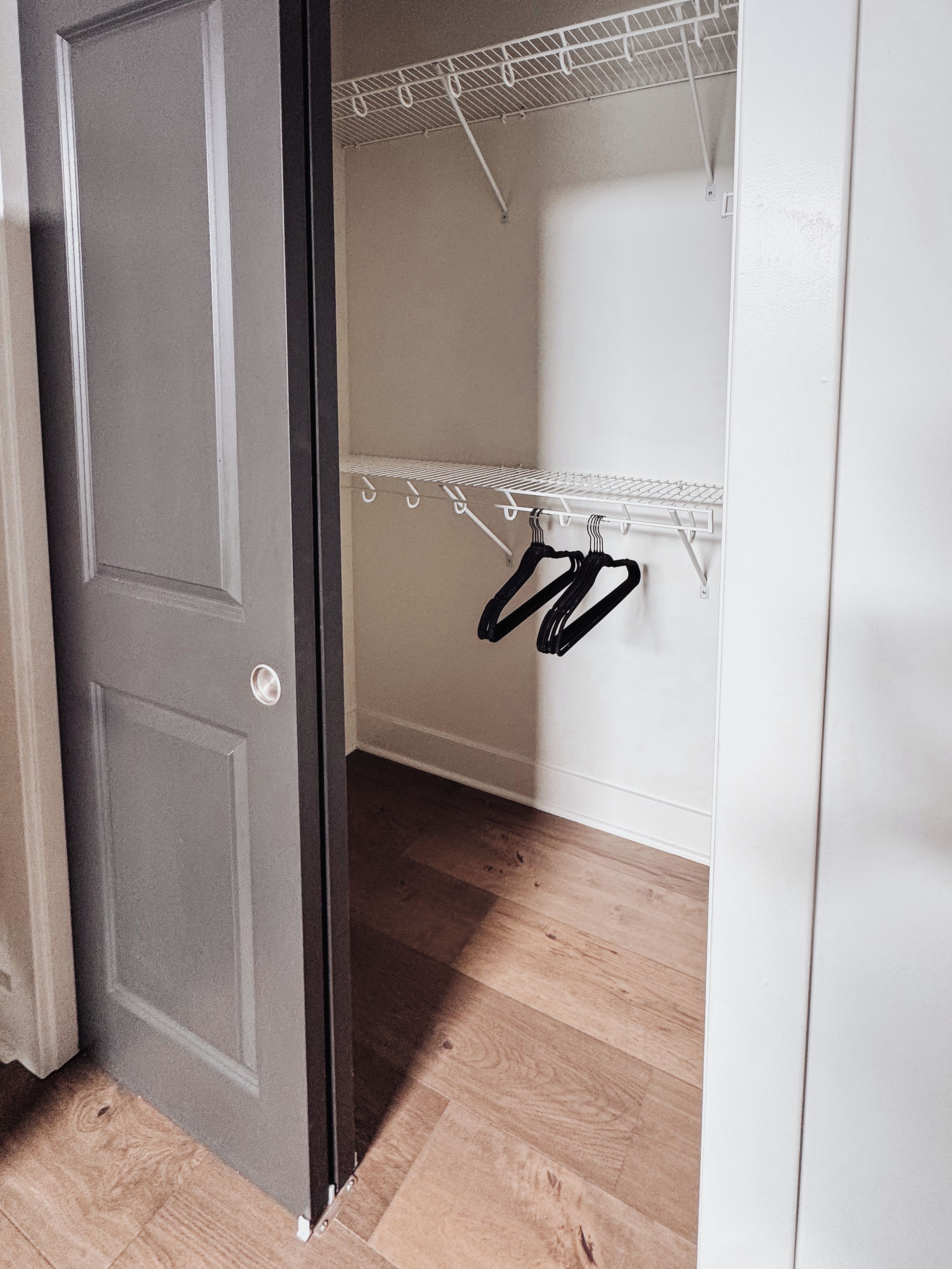 Una habitación con piso de madera, pared blanca y una puerta que da a un armario con estante metálico.