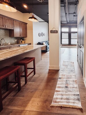 A modern kitchen with a marble countertop, wooden stools, and a hanging light fixture.