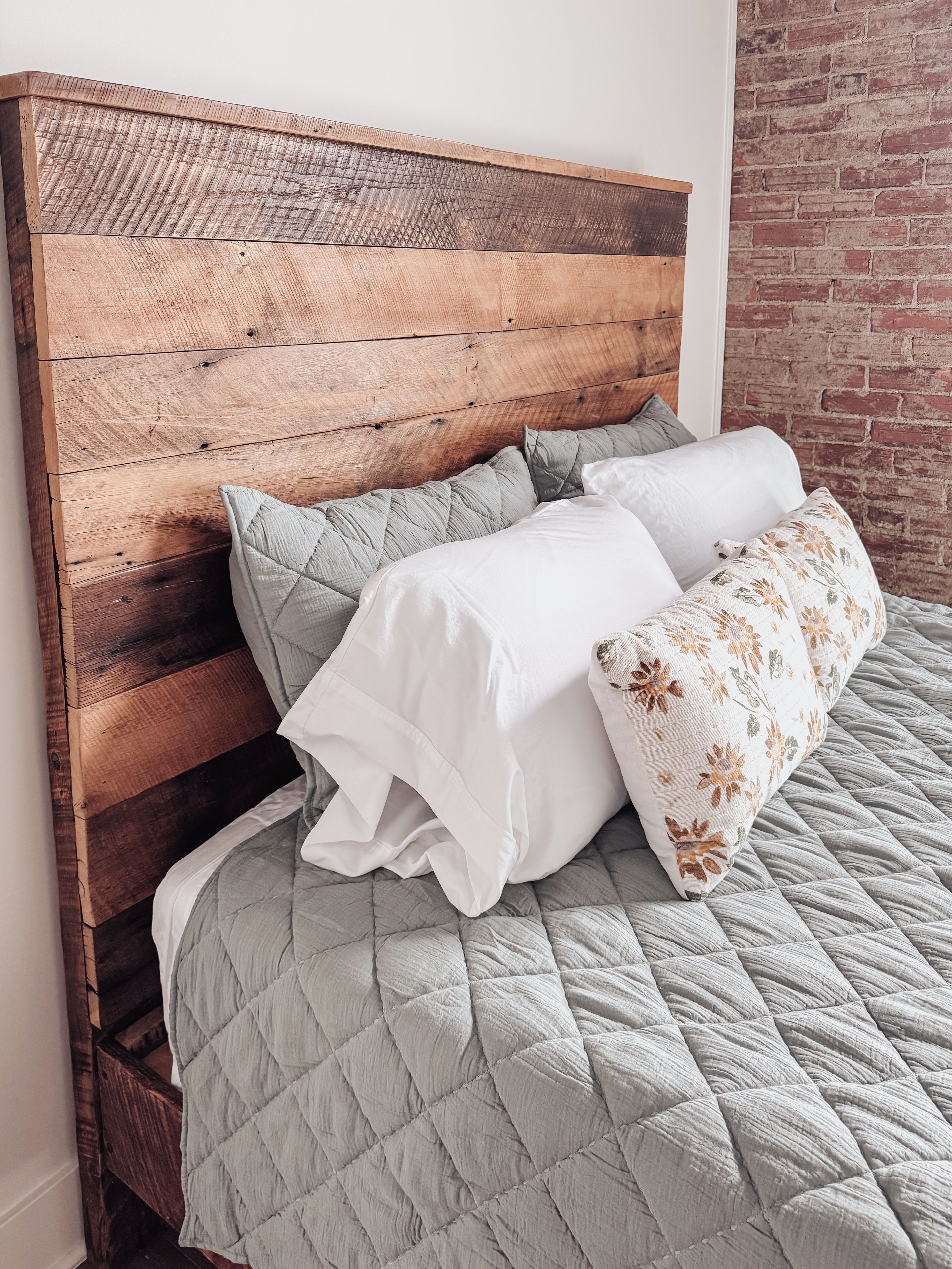 Una cama con cabecera de madera, cubrecama gris acolchada y dos almohadas.