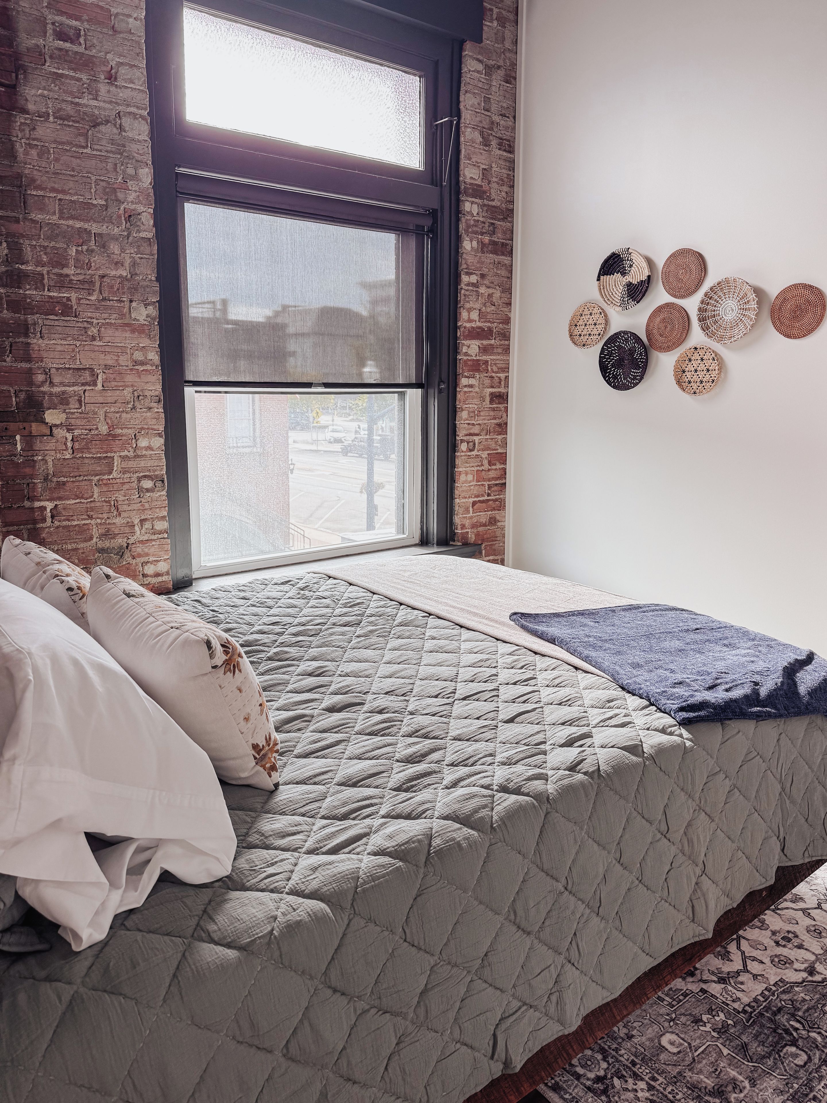 A room with a bed, a window with blinds, a brick wall, and a wall with circular wall hangings.