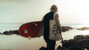 A person carrying a surfboard on a beach with rocky terrain and the ocean in the background. - Agachill Surf House (Aourir)