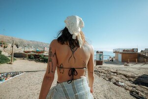 Escena de playa con una persona usando un pañuelo y un traje de baño, una costa rocosa y un edificio al fondo.