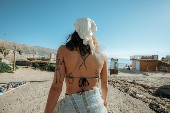 A beach scene with a person wearing a headscarf and a bikini, with a rocky shoreline and a building in the background. - Agachill Surf House (Aourir)