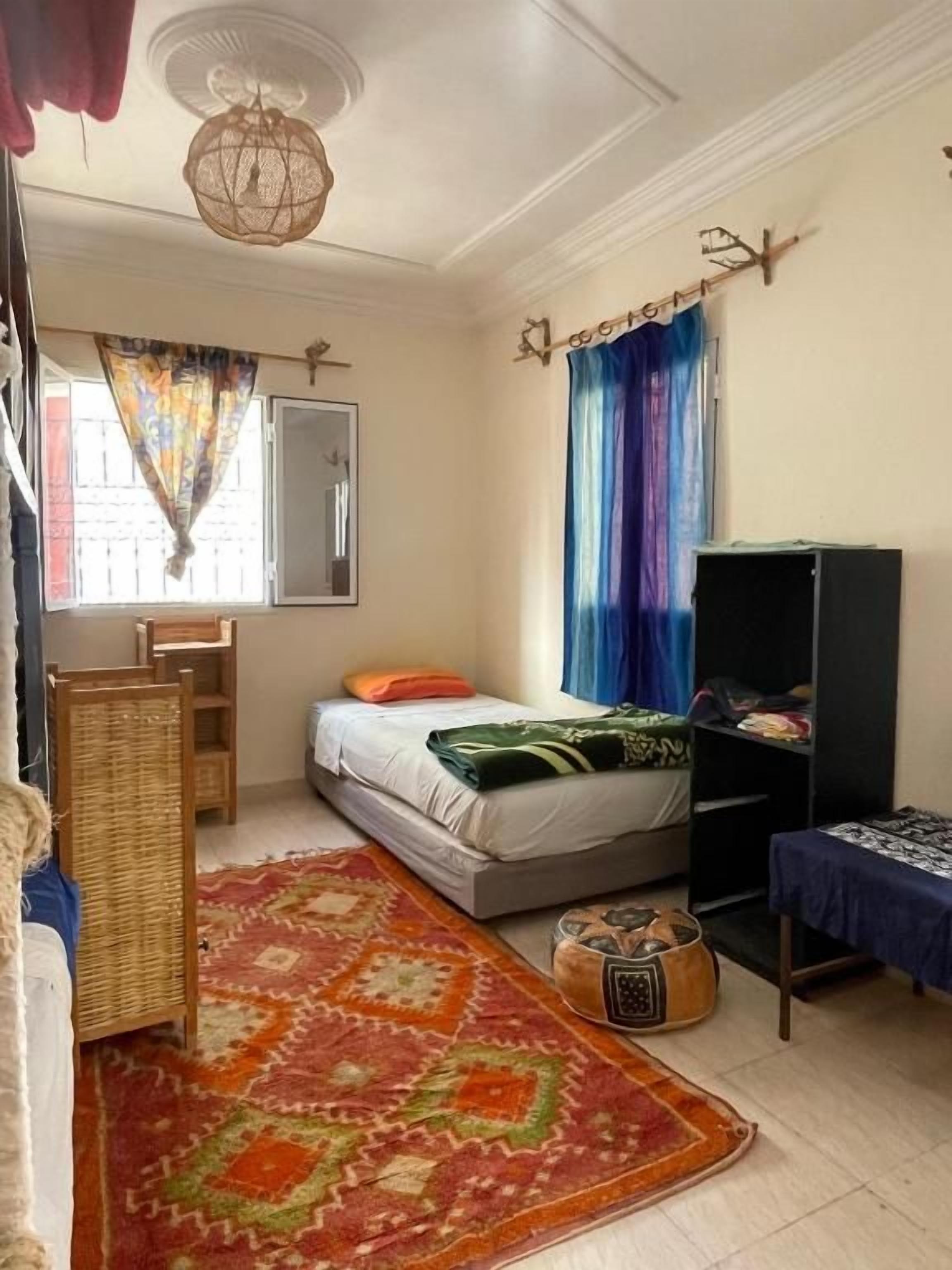 A bedroom with a bed, a wardrobe, a desk, a chair, a rug, and a window with curtains.