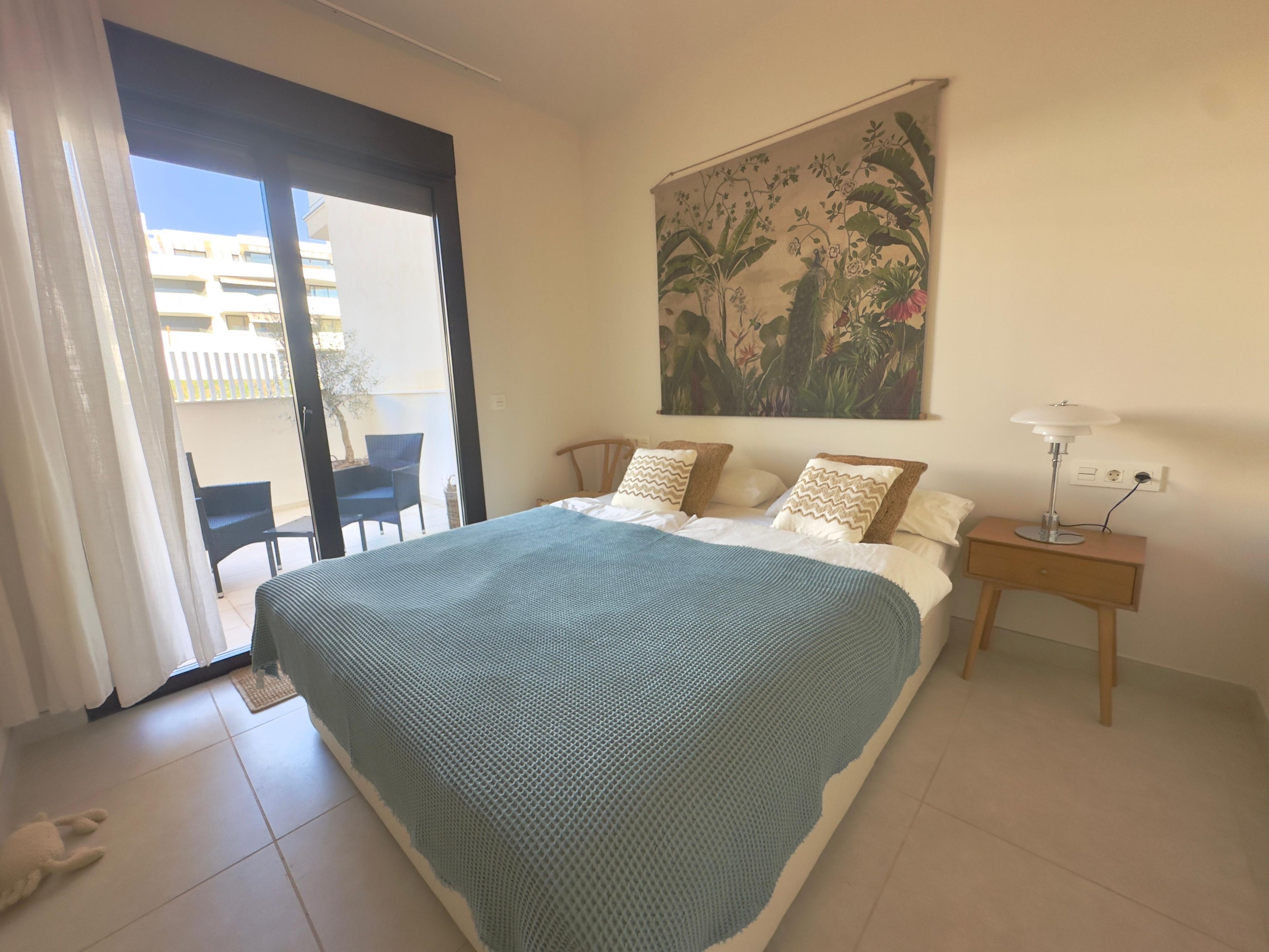 A bedroom with a bed, a nightstand, a lamp, a painting, a chair, and a view of the outside.