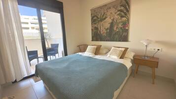 A bedroom with a bed, a nightstand, a lamp, a painting, a chair, and a view of the outside.