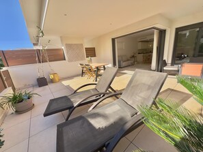 Un espacio al aire libre iluminado por el sol con sillones de descanso y una mesa, una puerta corrediza de vidrio que da a un espacio interior, y plantas en macetas.