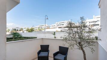 A balcony with two wicker chairs, a potted plant, and a view of a white fence and buildings.