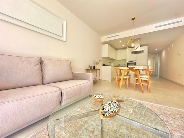 Una sala moderna con un sofá, una mesa de centro de vidrio con adornos, una zona de comedor con sillas de madera y una cocina al fondo.