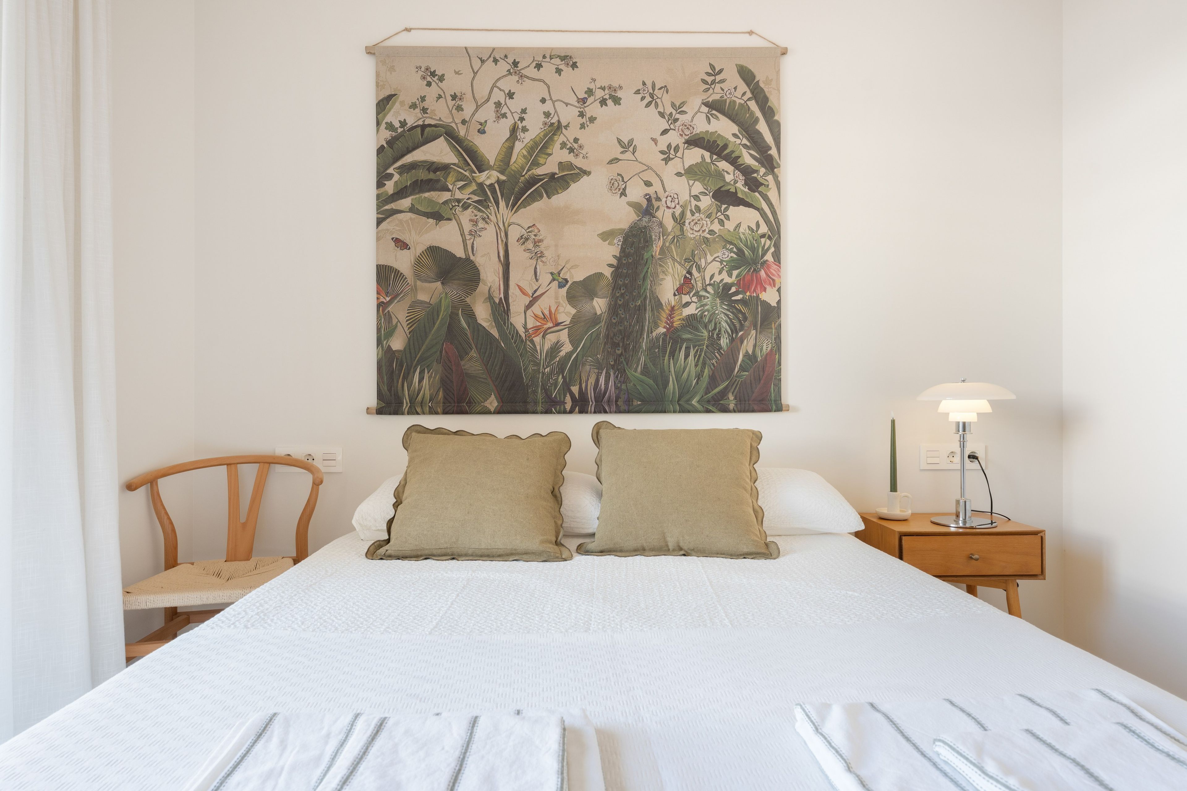 A bedroom with a bed, two pillows, a nightstand with a lamp, a chair, and a wall hanging.