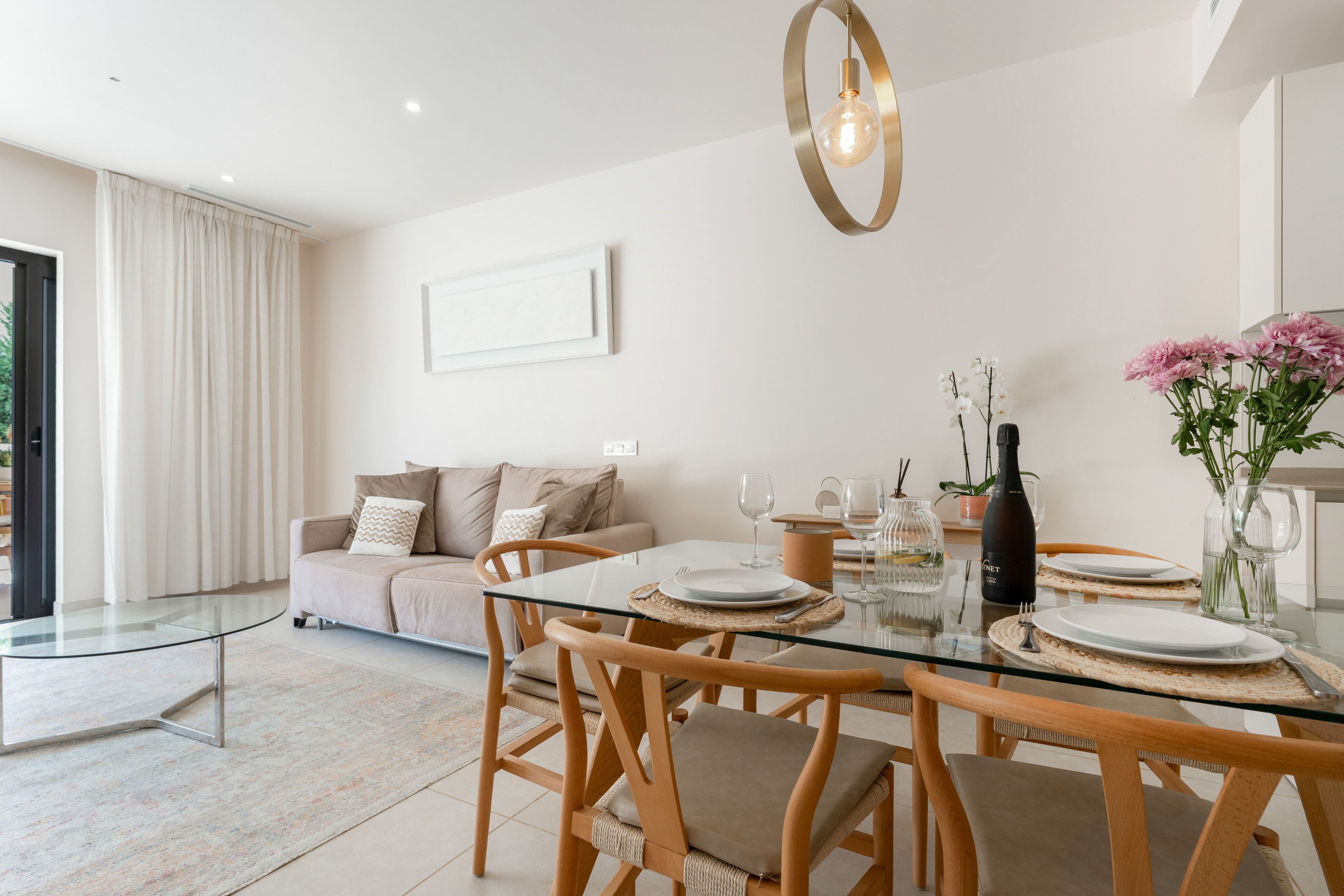 A modern dining area with a glass table, wooden chairs, and a sofa in the background.