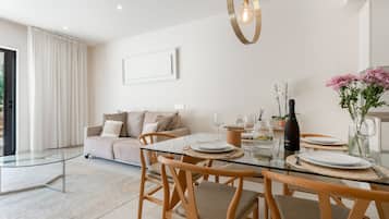 A modern dining area with a glass table, wooden chairs, and a sofa in the background.