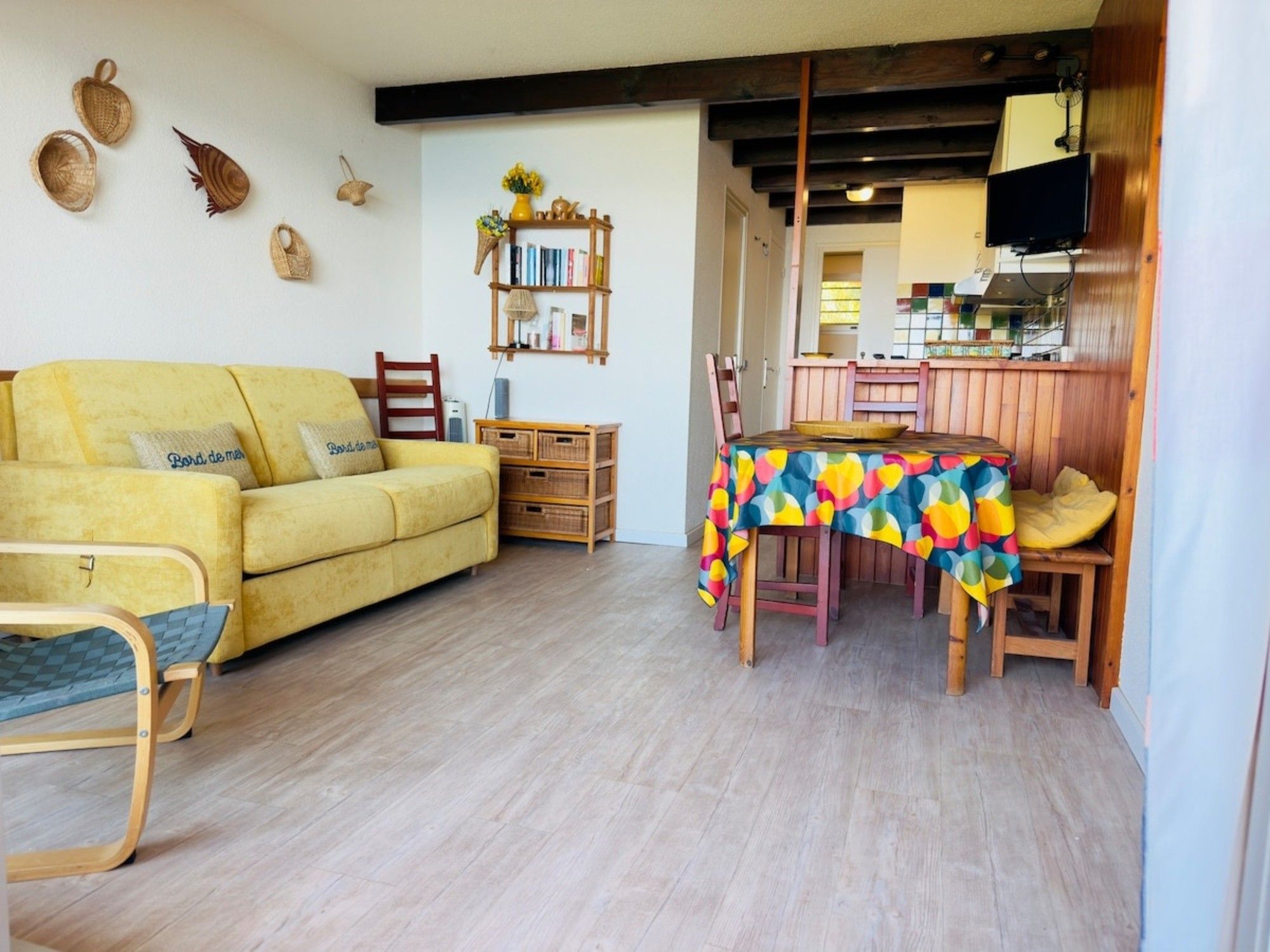 A living room with a yellow sofa, a wooden dining table with a colorful tablecloth, and a kitchen area in the background.