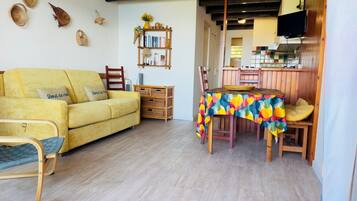 A living room with a yellow sofa, a wooden dining table with a colorful tablecloth, and a kitchen area in the background.
