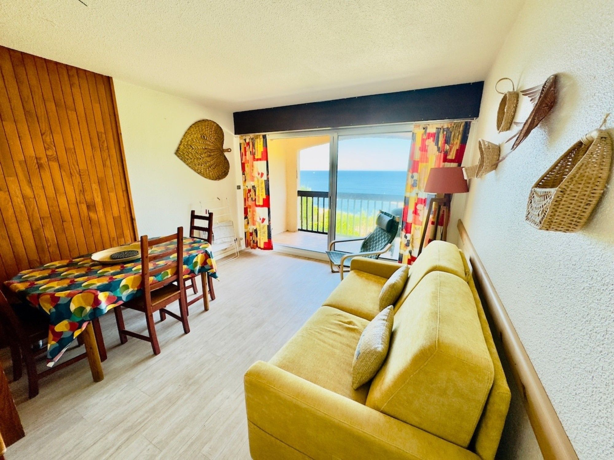 A room with a wooden dining table, a yellow sofa, a view of the sea through a sliding glass door, and a patterned wall hanging.