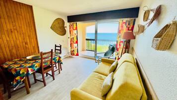 A room with a wooden dining table, a yellow sofa, a view of the sea through a sliding glass door, and a patterned wall hanging.