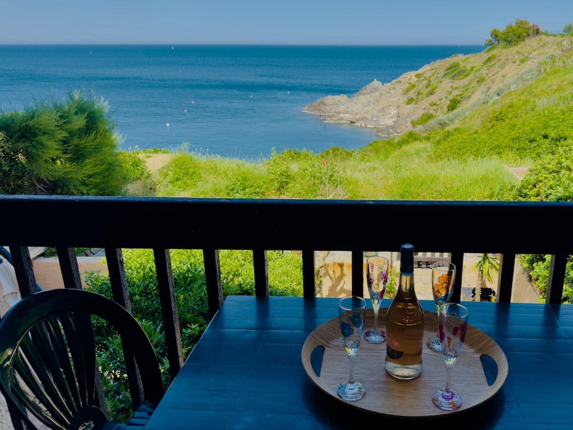 Ein Tisch mit einer Flasche und Gläsern, mit Blick auf eine Küstenlandschaft mit Meer und Hügeln.