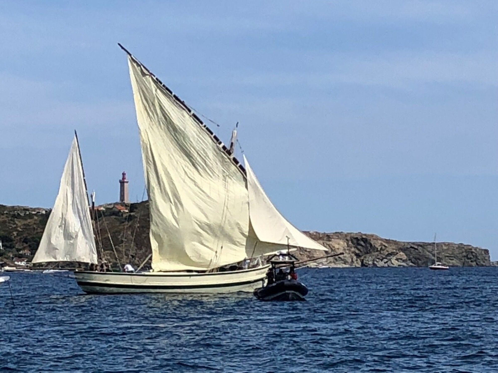 En segelbåt med ett stort segel på vattnet, en klippig kustlinje och en fyr i bakgrunden.