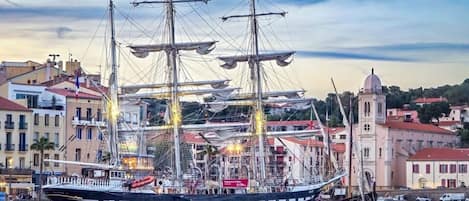 A large sailing ship docked at a harbor with buildings and a tower in the background.