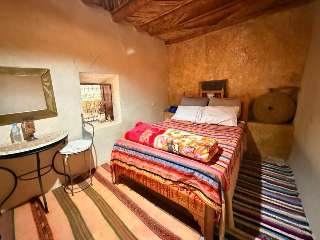A bedroom with a bed, a small table with a chair, a window, and a wooden ceiling.