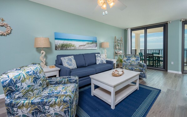 A living room with a sofa, armchairs, a coffee table, and a painting of a beach scene. - Phoenix East 308 1 Bedroom Condo (Orange Beach)