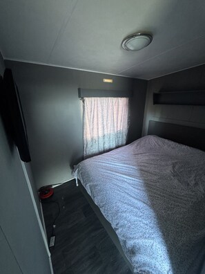 Ein kleines Zimmer mit einem Bett, einem Vorhangfenster, einer Deckenleuchte und einem an der Wand montierten Fernseher.