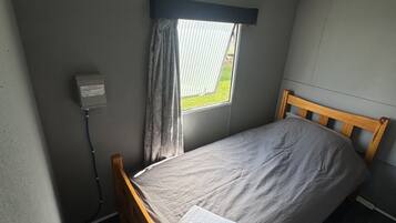 Una cama individual con cabecera de madera en una habitación pequeña con ventana y una lámpara fijada a la pared.