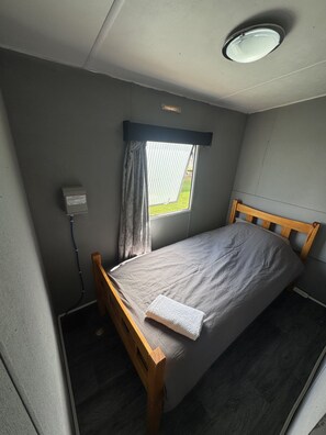 A single bed with a wooden headboard in a small room with a window and a wall-mounted light fixture.