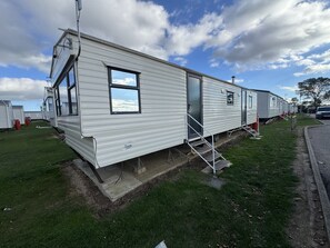 Ein Mobilheim mit einer Veranda und einem Fenster, gelegen auf einer Rasenfläche, im Hintergrund sind weitere ähnliche Häuser zu sehen.