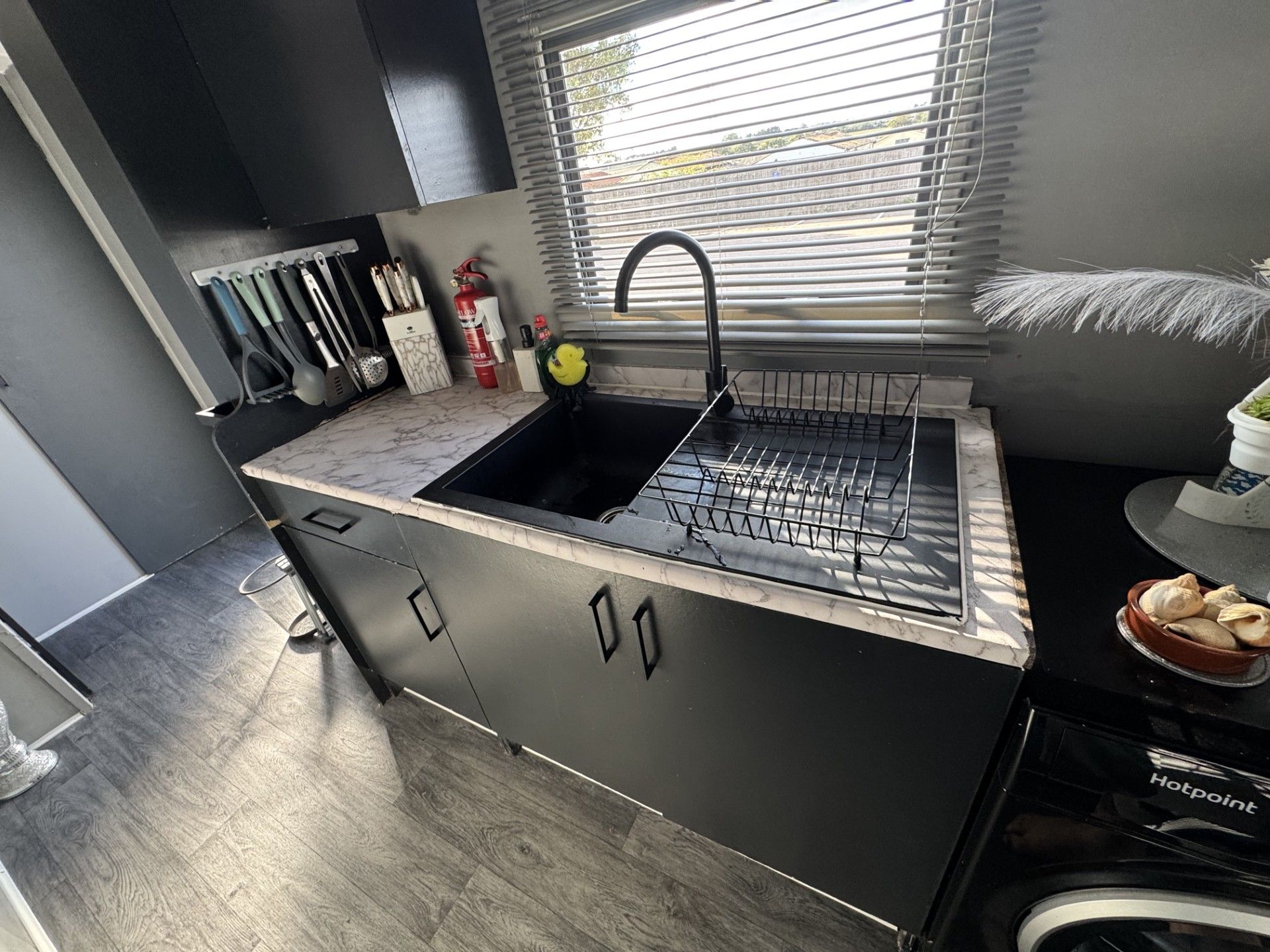A compact kitchen with a black sink, a window with blinds, and a dish rack.