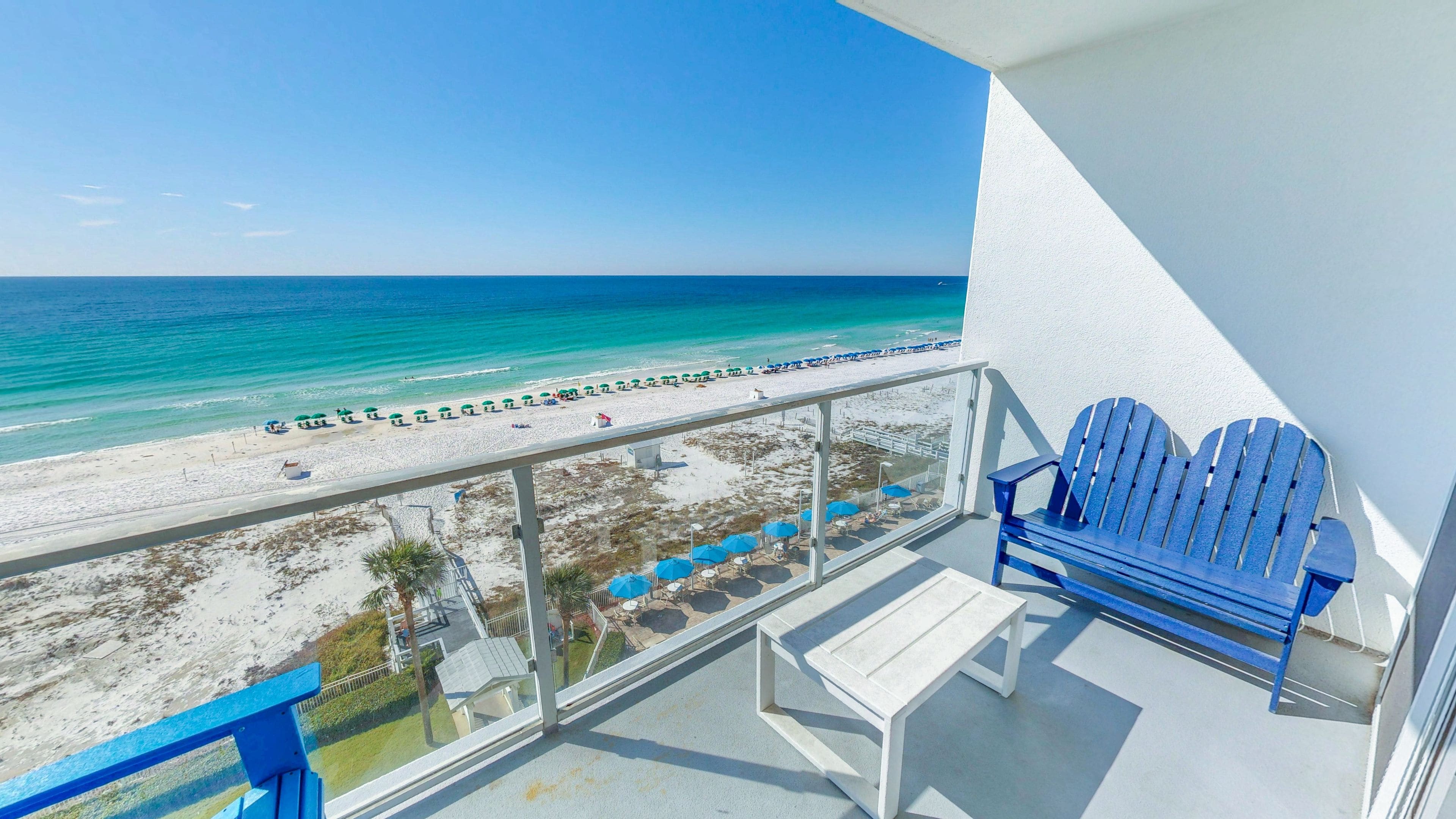 A balcony with a view of a beach and ocean. There are two blue Adirondack chairs and a white table.