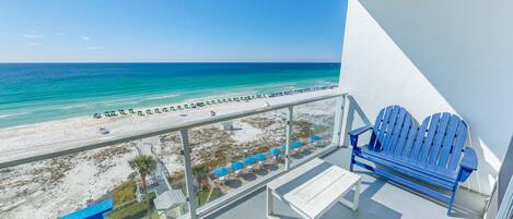 A balcony with a view of a beach and ocean. There are two blue Adirondack chairs and a white table.