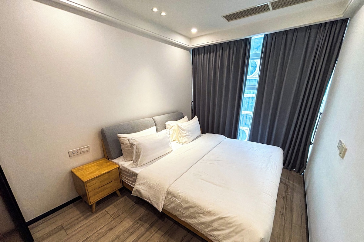 A modern hotel room with a large bed, a wooden nightstand, and a window with curtains.