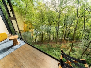 Ein Zimmer mit großem Fenster mit Blick auf einen Wald. Es gibt eine hölzerne Bank und einen kleinen Hocker.
