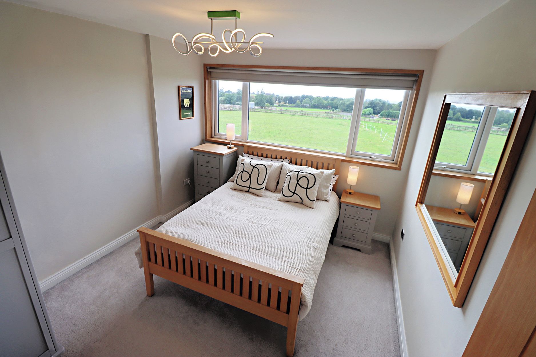 Une chambre à coucher avec un lit en bois, des tables de chevet, une commode, une fenêtre donnant sur un champ verdoyant et un miroir.