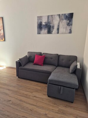 A sectional sofa with a red accent pillow, a small ottoman, and a wall-mounted abstract art piece. - Floor 10 - Tenth Floor (Roma)
