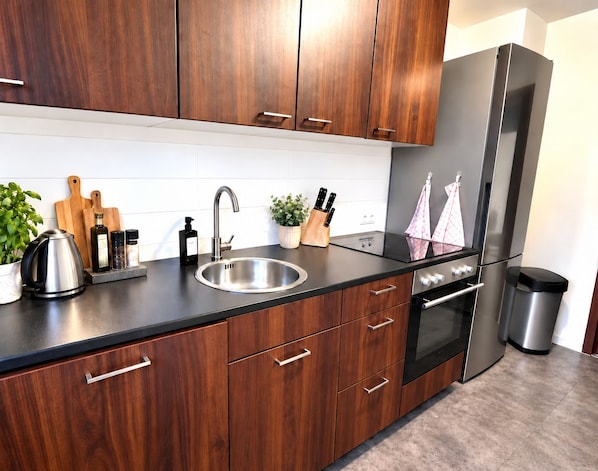 A modern kitchen with wooden cabinets, a stainless steel sink, a black countertop, a built-in oven, and a stainless steel refrigerator.