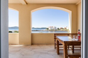 A balcony with a view of a body of water and buildings, a wooden table, and a bottle of water. - Kotari Sukošan in Sukošan - Nord-Dalmatien / Kotari Sukošan in Sukošan - Appartement für 4 Personen AP5 (Sukošan)