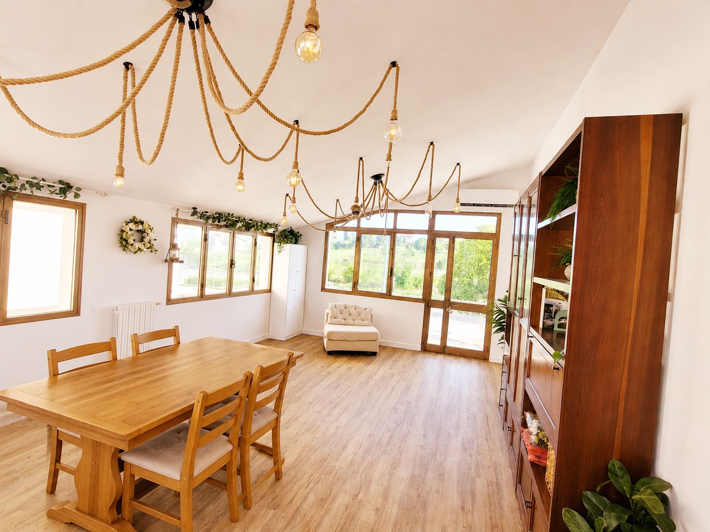 A dining area with a wooden table and chairs, a chandelier, and large windows with a view of greenery.
