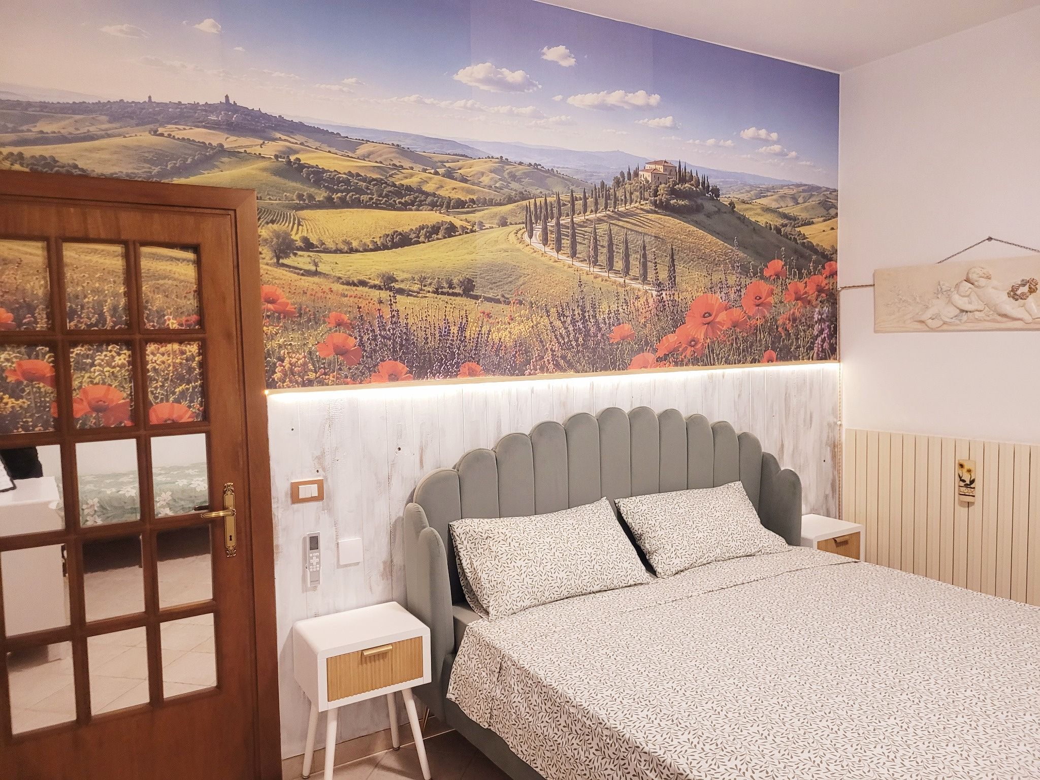 Una camera da letto con un letto, un comodino, una porta e un affresco raffigurante un paesaggio.