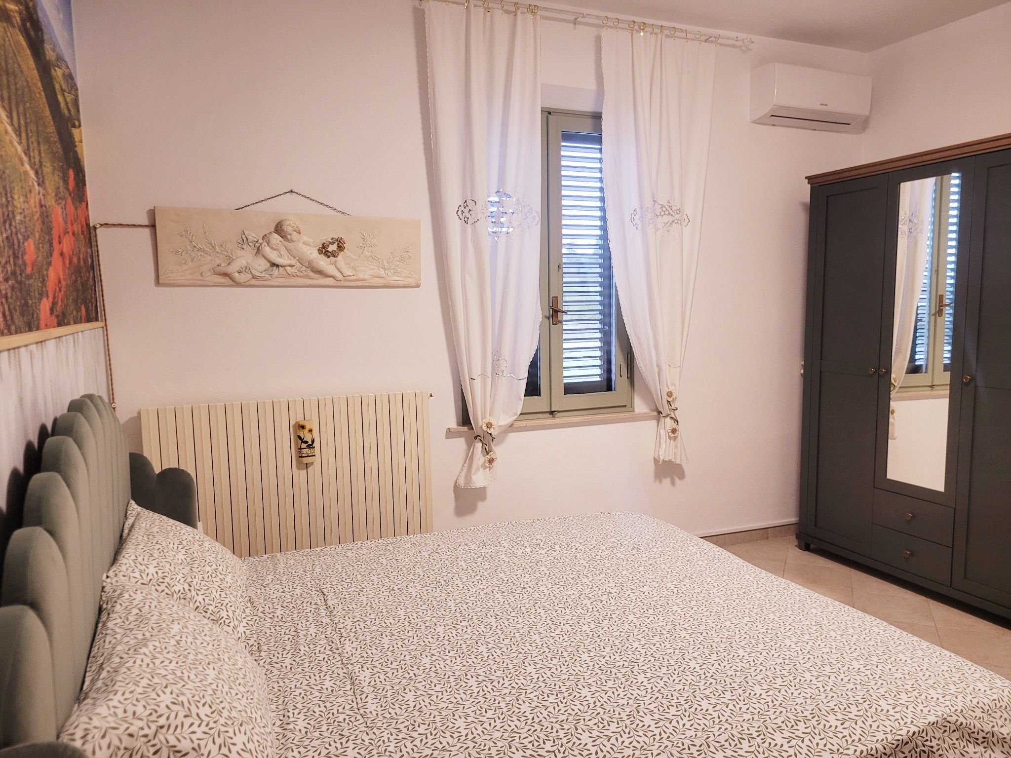 A bedroom with a bed, a radiator, a window with curtains, a wall-mounted air conditioner, and a wardrobe.